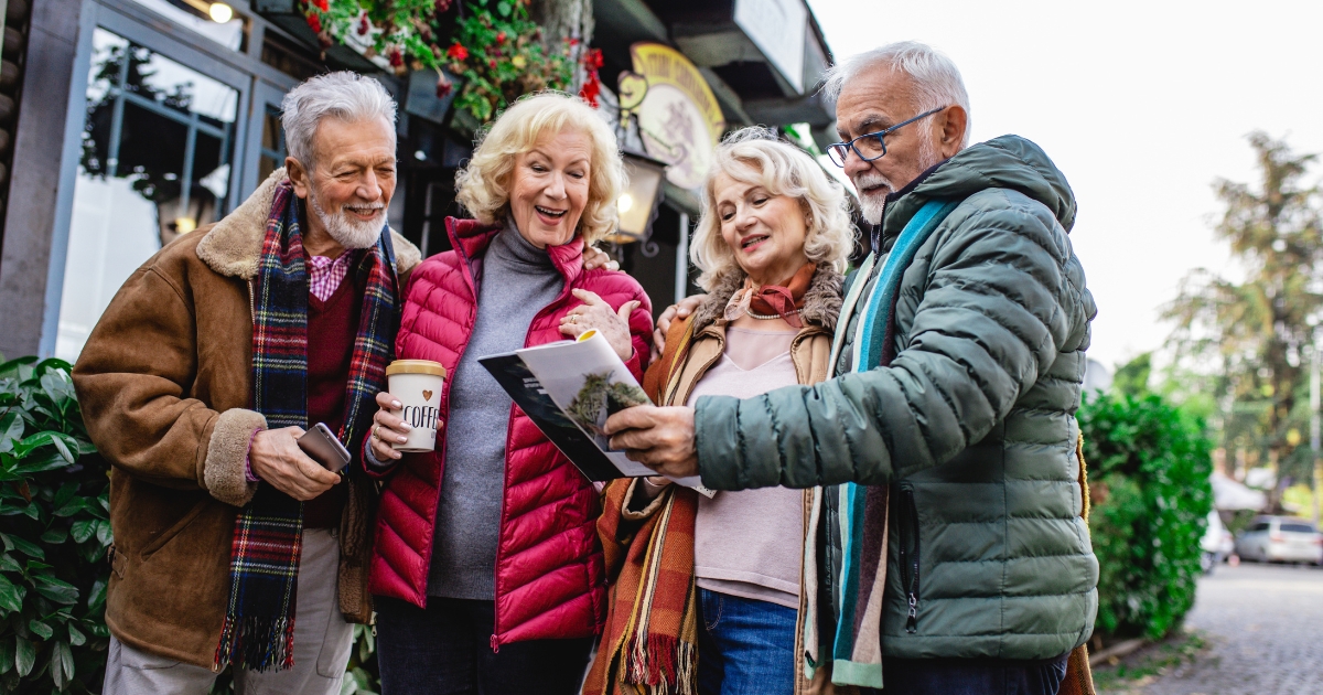 Group of happy seniors bundled in winter clothing, smiling and looking at a map or brochure outdoors — representing active socializing and travel for people over 60.
