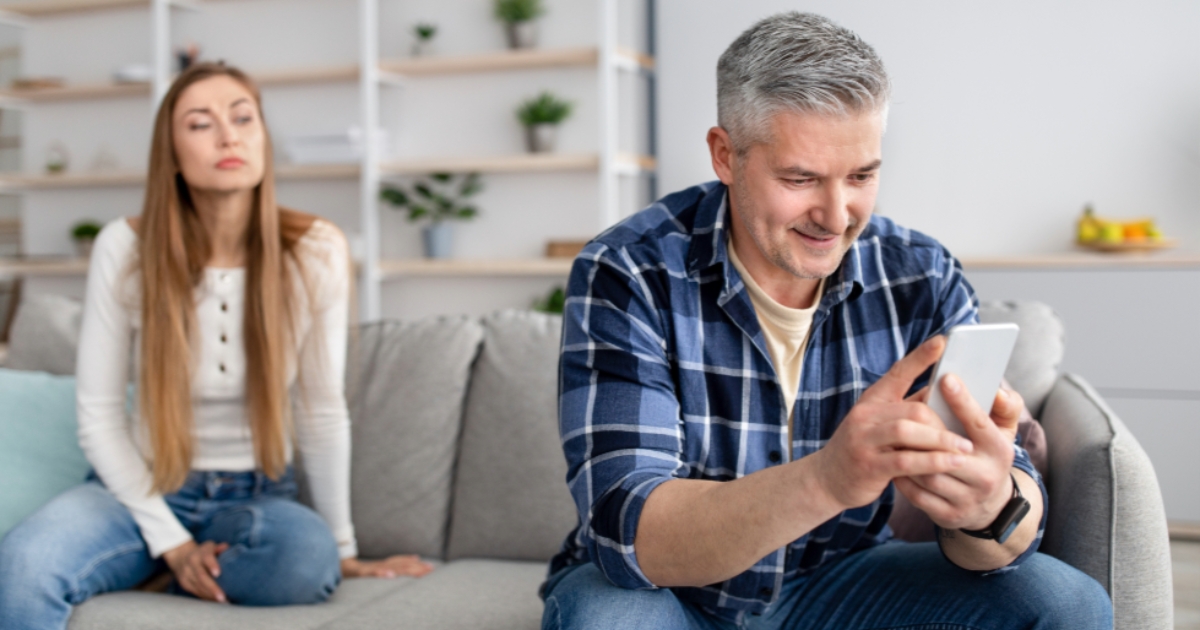Older man smiling while texting on his phone, seated next to a younger woman looking upset—illustrating relationship tension and signs of emotional disconnection or possible infidelity.