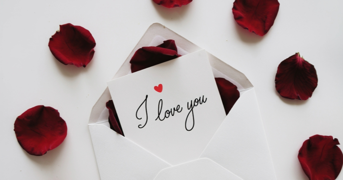 A white envelope with a card inside that reads 'I love you' with a small red heart above the text, surrounded by red rose petals on a white background.