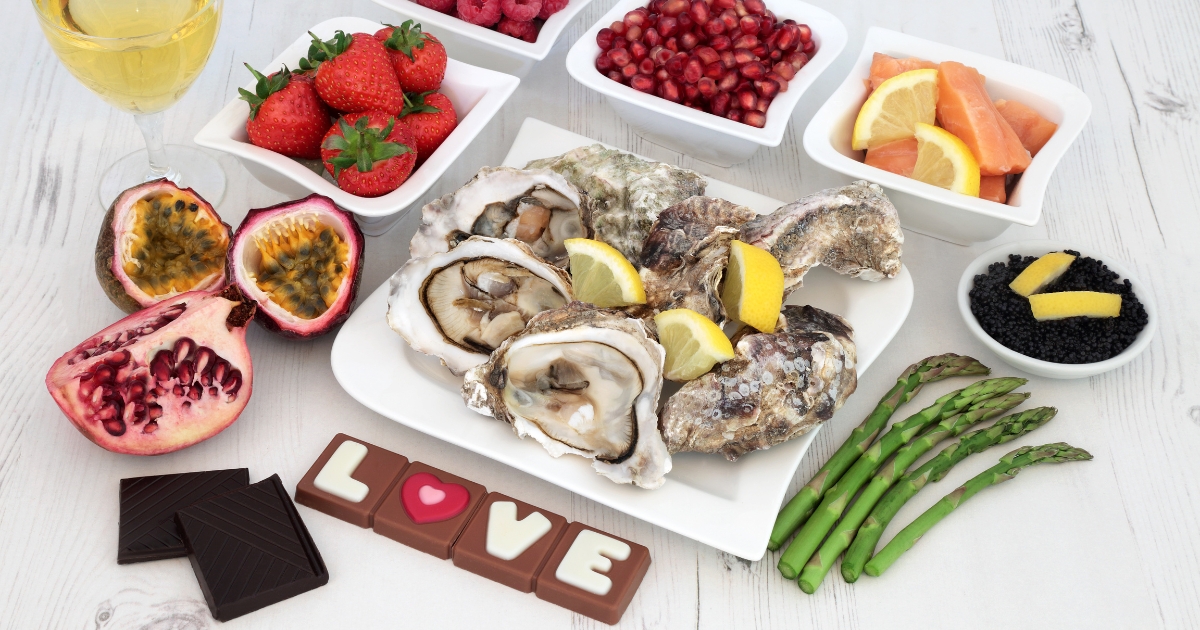 Aphrodisiac foods including oysters, dark chocolate, strawberries, pomegranate, asparagus, passion fruit, and salmon arranged on a white table with “LOVE” chocolates — symbolizing romance, libido-boosting nutrition, and sensual dining.