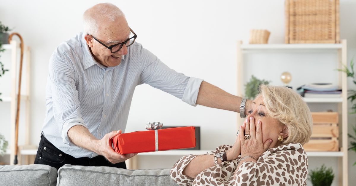 5 Simple Gestures For Saying I Love You Without Uttering A Word - Mature Singles Finding Love Happy elderly man surprising his wife with a red gift box, while she reacts with excitement and joy in a cozy living room setting.