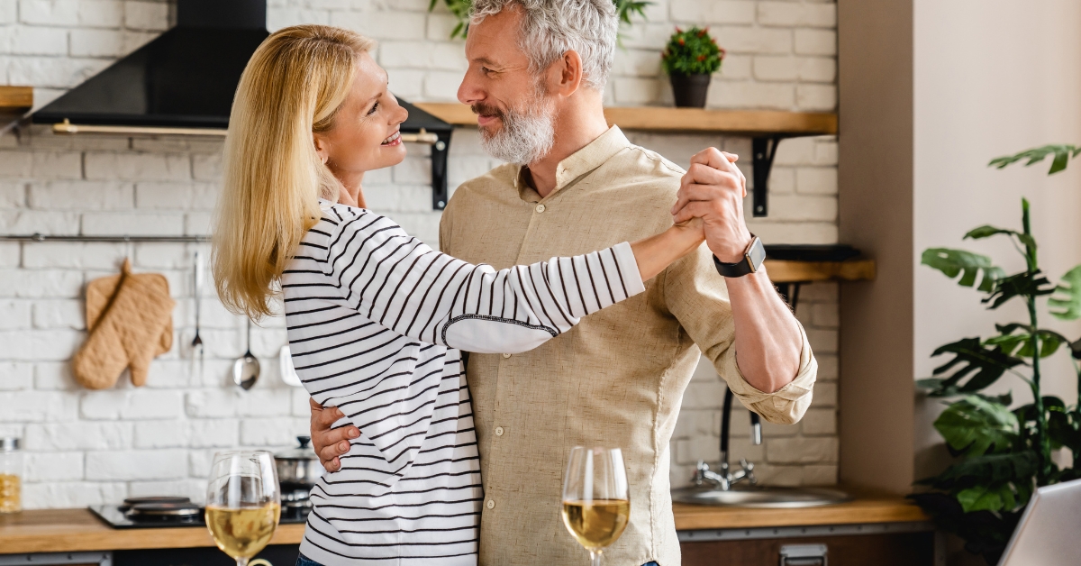 8 Simple Ways To Improve Your Sex Life In Your 50s - Mature Singles Finding Love Happy middle-aged couple dancing together in a cozy kitchen, enjoying a romantic moment with glasses of white wine on the counter. A warm and intimate setting highlighting love, connection, and joy in relationships over 50.