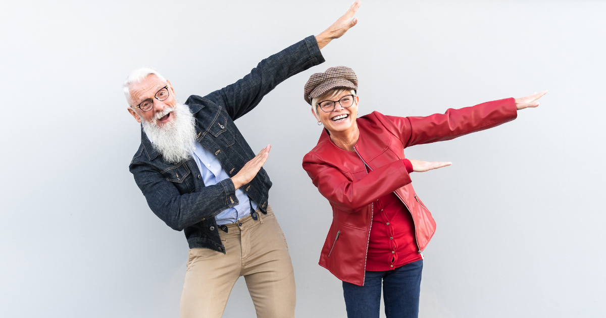 Age is Just a Number_ Dispelling Myths About Love in Later Life - Mature Singles Finding Love An energetic elderly couple striking fun poses with big smiles. The man has a white beard and is wearing glasses, a denim jacket, and beige pants, while the woman is wearing glasses, a stylish cap, a red jacket, and a matching top. Both are radiating joy and vitality against a neutral background.