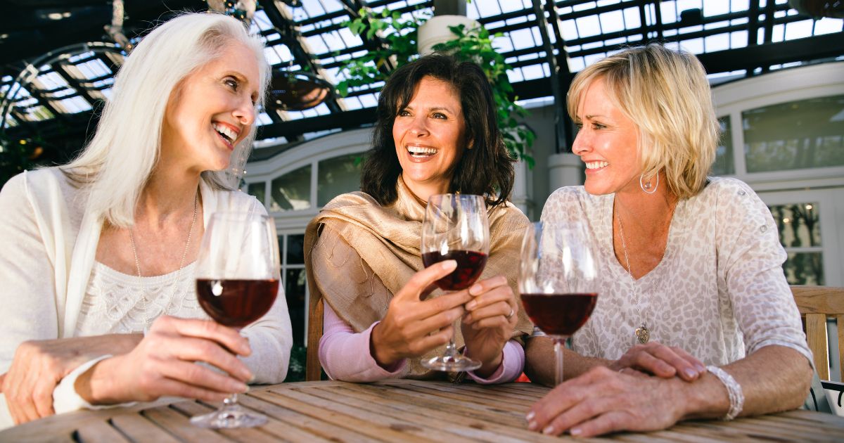 Three middle-aged women laughing and enjoying red wine together at an outdoor café.