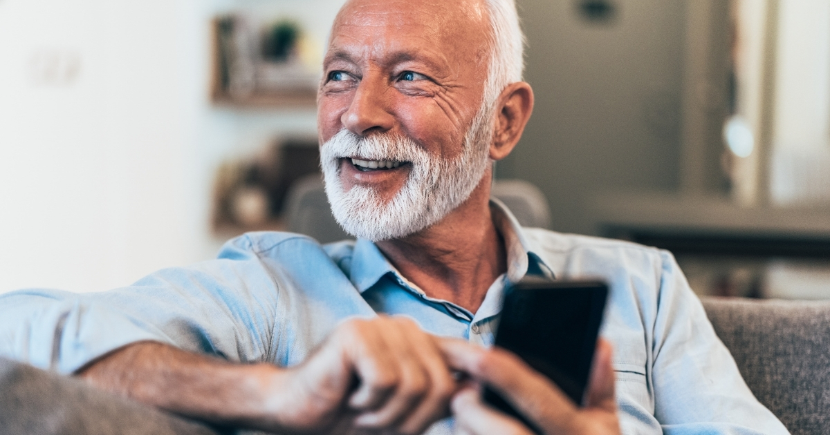 Are You Texting Too Much_ How To Know If You Are Coming On Too Strong - Mature Singles Finding Love Smiling elderly man with a white beard sitting on a couch, holding a smartphone, appearing relaxed and happy.