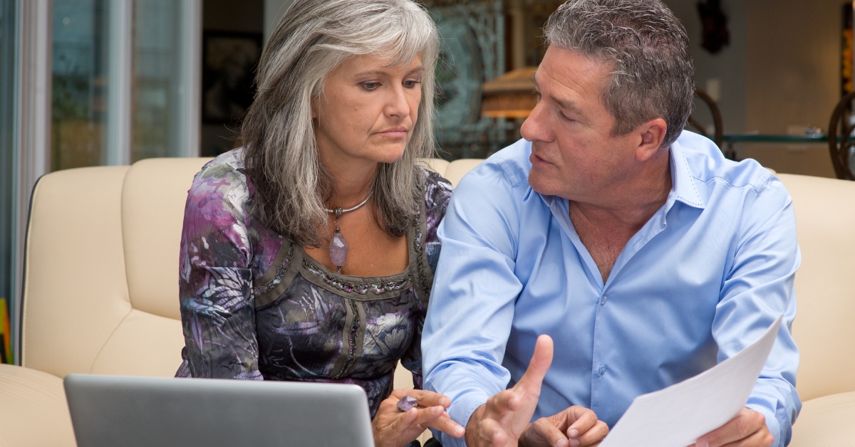 Avoiding Common Pitfalls in Senior Dating - Mature Singles Finding Love A mature couple sitting on a couch, engaged in a serious conversation while looking at a laptop and holding a document. The woman appears concerned, while the man is explaining something, suggesting a discussion about finances, online matters, or relationship concerns.