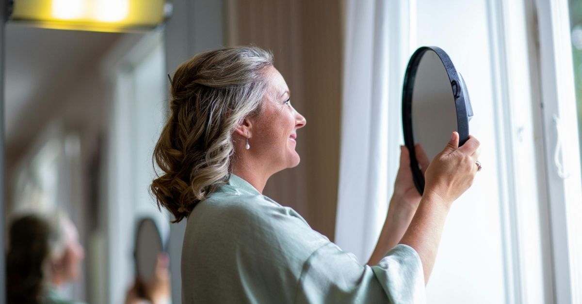 Mature woman with graying hair smiling while holding a round mirror near a window, symbolizing self-reflection, personal growth, and empowerment.