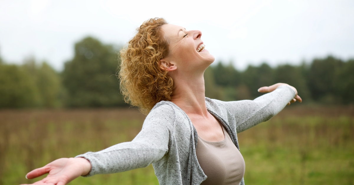 Dating After Divorce_ Hope and Healing in Your 50s - Mature Singles Finding Love A joyful woman with curly hair standing in an open field, arms outstretched, smiling with her face tilted upwards, embracing nature and freedom.