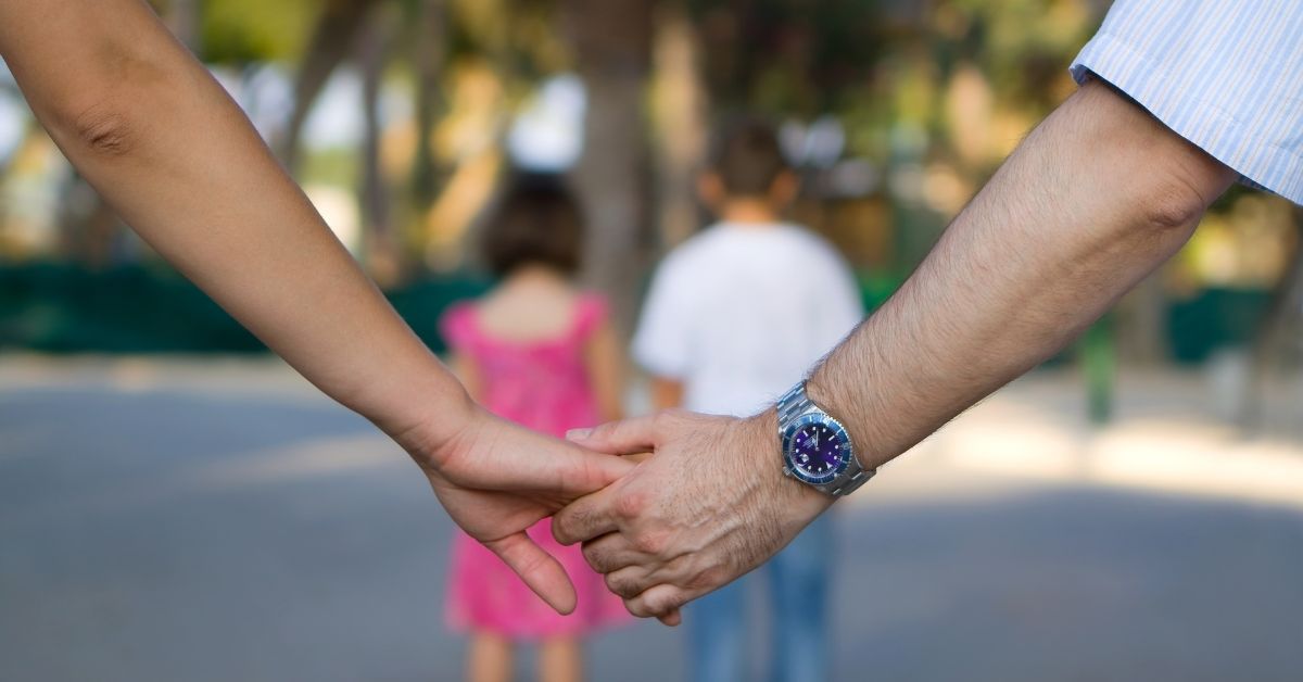 Dating Tips For Single Parents_ How To Include The Kids - Mature Singles Finding Love Two adults holding hands in the foreground with two children standing in the background, outdoors in a park setting.