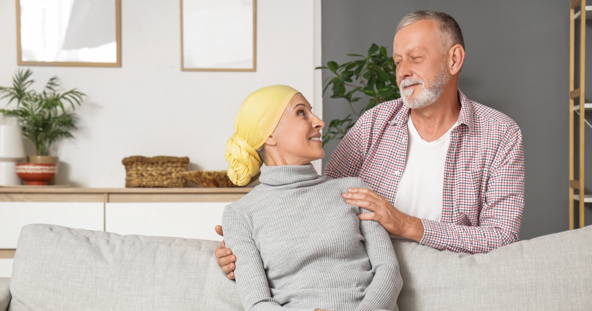 Supportive senior couple sitting together on a couch, with the woman wearing a yellow headscarf and smiling at her partner in a warm home setting.