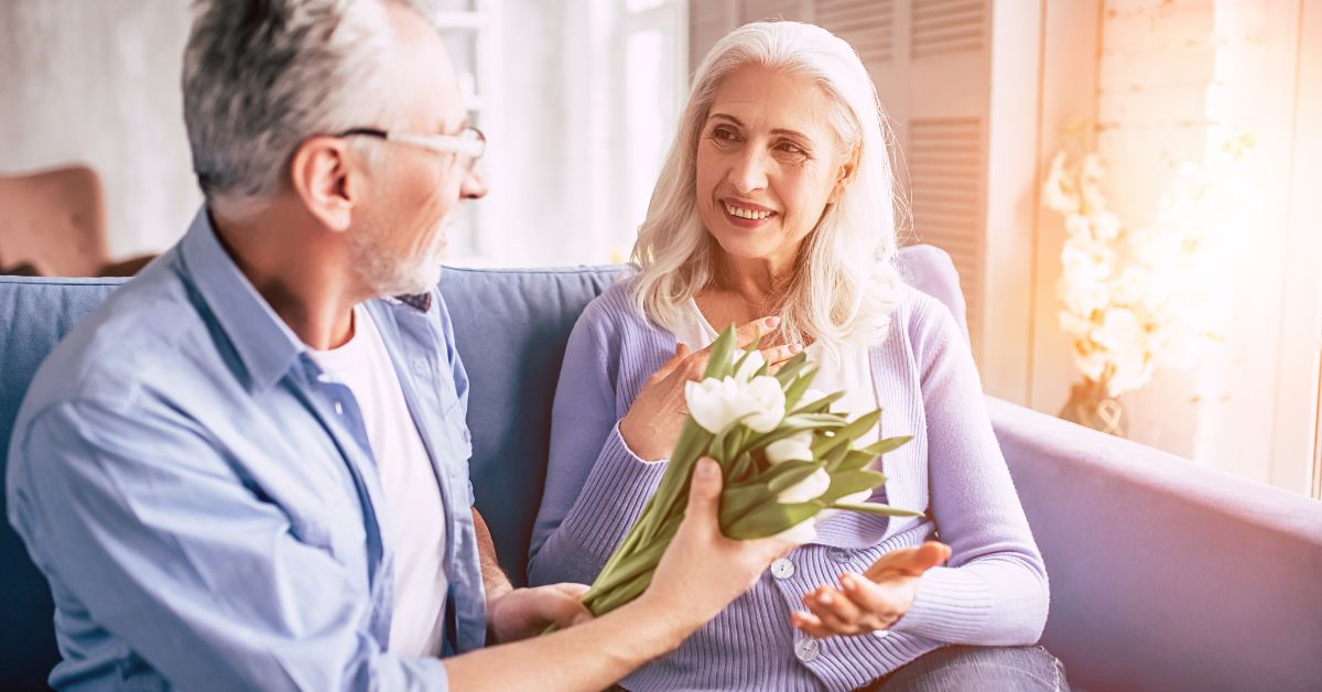 Do You Know Your Love Language_ How To Find Out - Mature Singles Finding Love Smiling older couple sitting on a couch, with the man giving white tulips to the woman as they share a heartfelt moment.