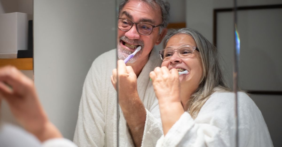 Happy older couple brushing teeth together in bathroom mirror, showing intimacy and daily connection after 50