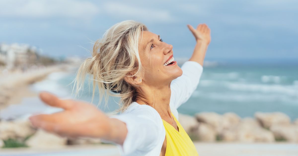 How Long Does It Take To Get Over A Divorce - Mature Singles Finding Love Joyful middle-aged woman with outstretched arms smiling by the ocean, symbolizing emotional freedom and healing after divorce.