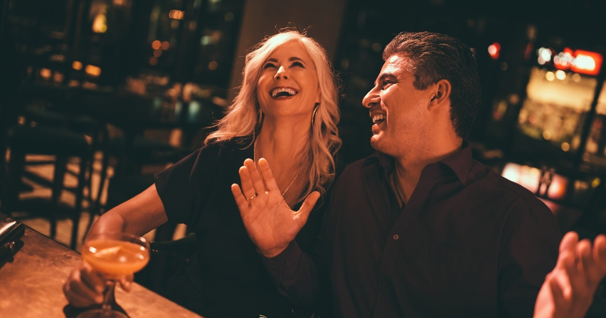 How Making A Woman Laugh Can Lead To A Relationship - Mature Singles Finding Love A couple sitting at a bar, laughing together while enjoying their drinks, showcasing a fun and lighthearted moment.