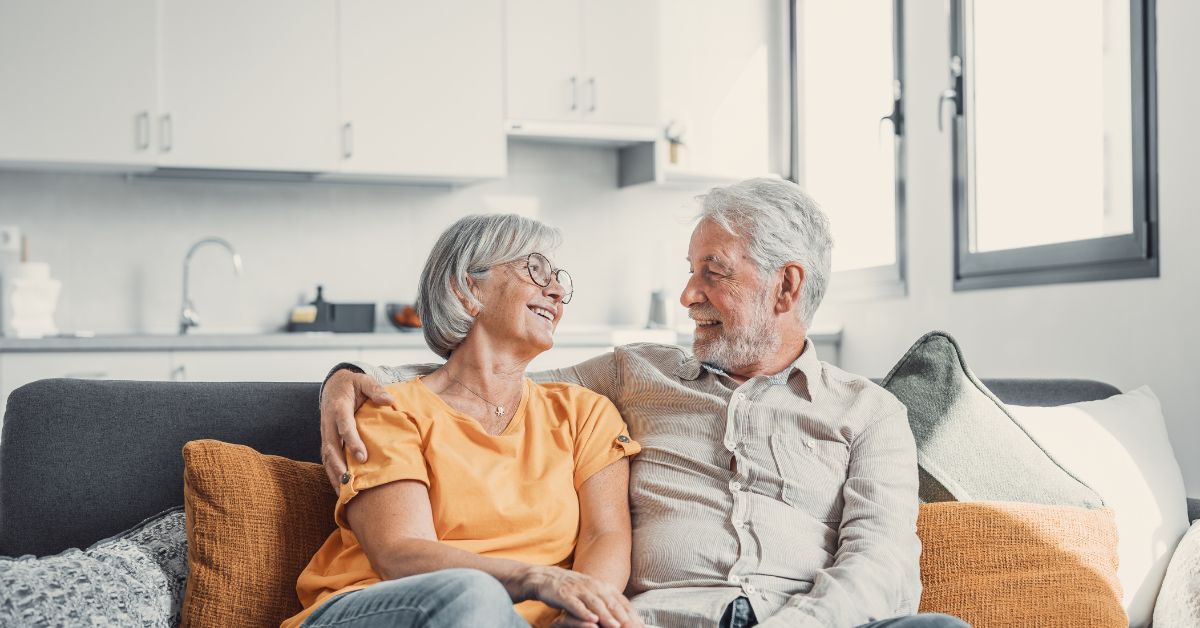 Happy senior couple sitting on a couch at home, smiling and looking at each other — symbolizing trust, emotional connection, and lasting love in mature relationships.
