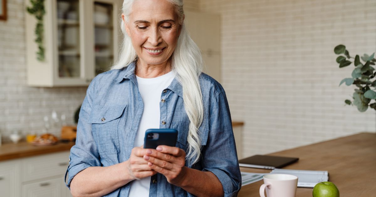 How To Make A Woman Feel Special Over Text - Mature Singles Finding Love Smiling older woman standing in a modern kitchen, looking at her smartphone while holding it with both hands. A coffee mug and a green apple sit on the counter.