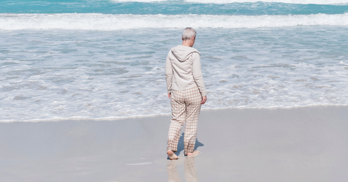 How To Move On After The Loss Of A Spouse - Mature Singles Finding Love An elderly person with short gray hair walking barefoot along the shoreline, wearing a beige hoodie and plaid pants, reflecting a serene and contemplative mood by the ocean.