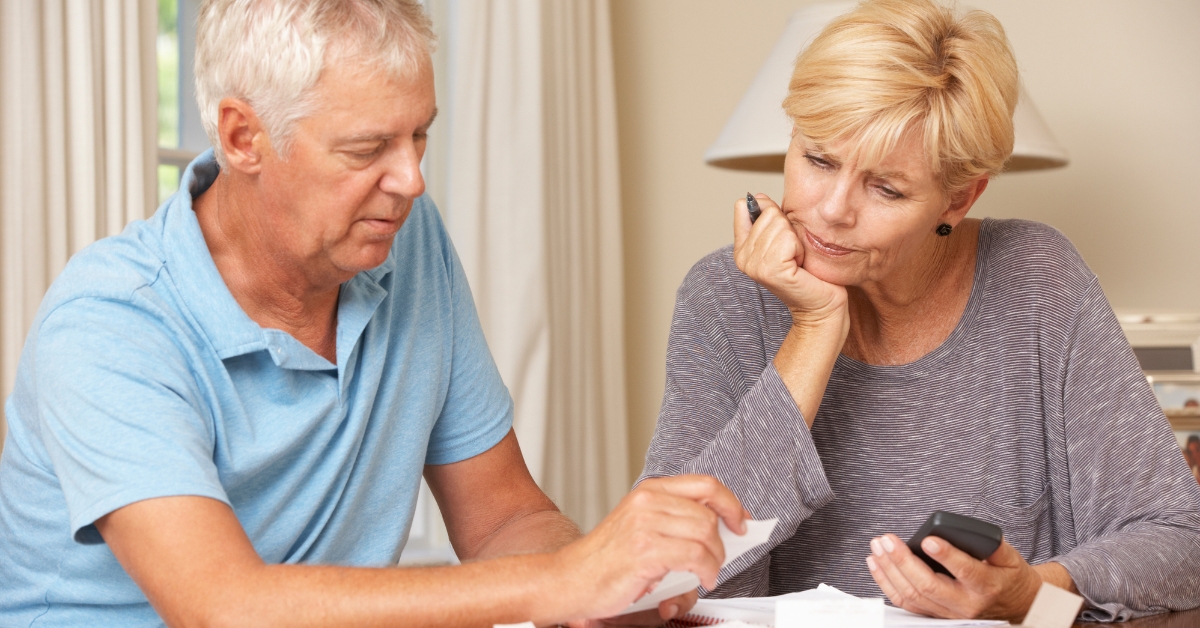 How to Discuss Financial Matters When Dating Over 50 - Mature Singles Finding Love A senior couple sitting at a table, reviewing financial documents together. The man in a blue polo shirt is holding a paper, while the woman in a gray top looks concerned, holding a calculator and a pen. They appear to be discussing financial matters in a home setting.