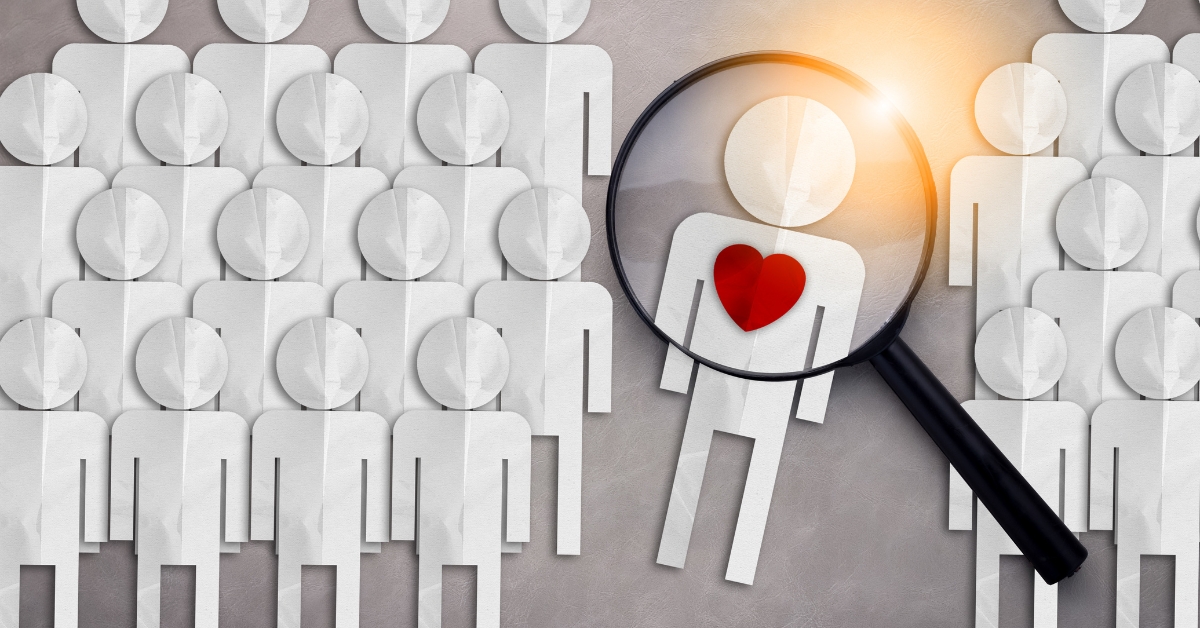 A magnifying glass highlighting a paper cutout figure with a red heart in a crowd of identical figures, symbolizing finding love or individuality in dating.