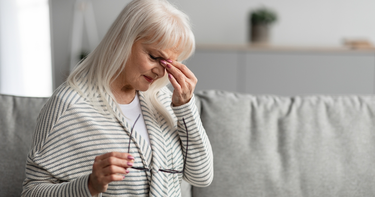 Senior woman sitting on a couch, holding her glasses and rubbing her eyes in frustration or sadness — representing emotional exhaustion or relationship stress.