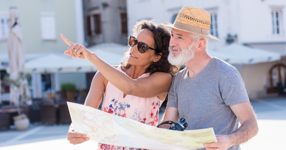 Happy senior couple exploring a city with a map, wearing casual summer clothes and sunglasses — representing travel, adventure, and active lifestyle for people over 60.