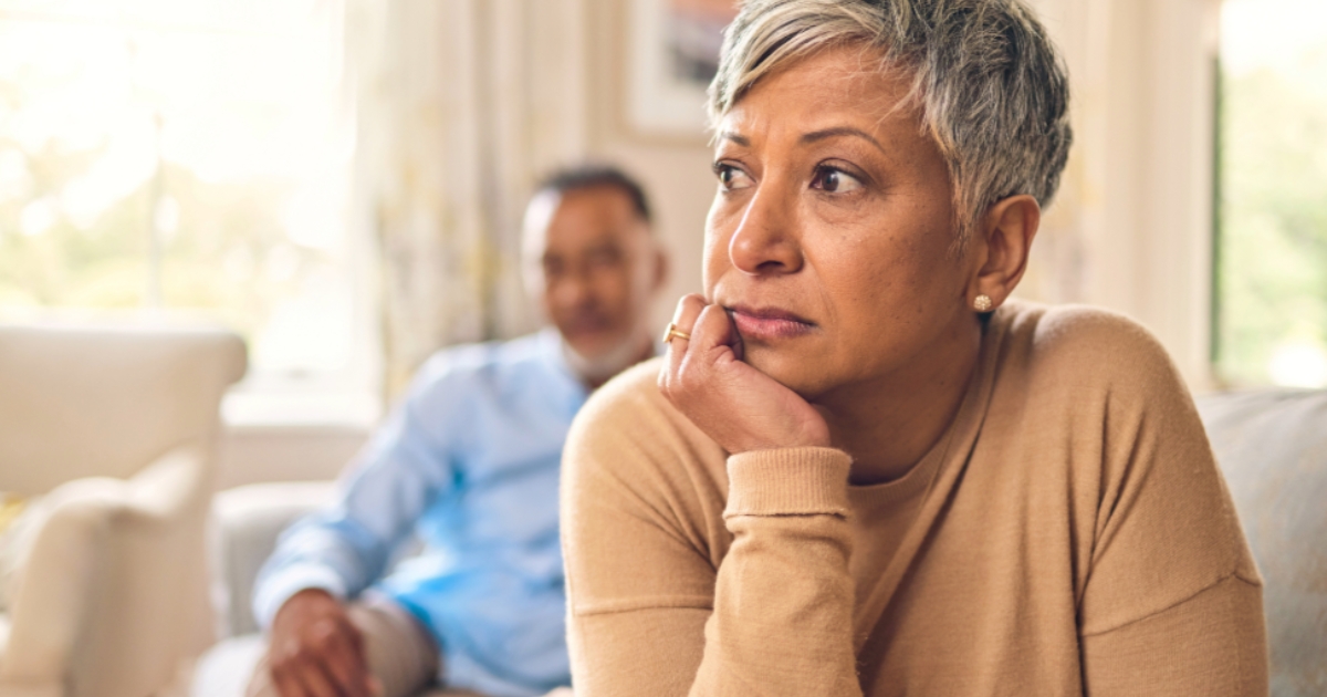 Upset mature woman sitting on a couch, looking away thoughtfully while a man sits in the background—symbolizing emotional distance, relationship conflict, or betrayal in a mature relationship.