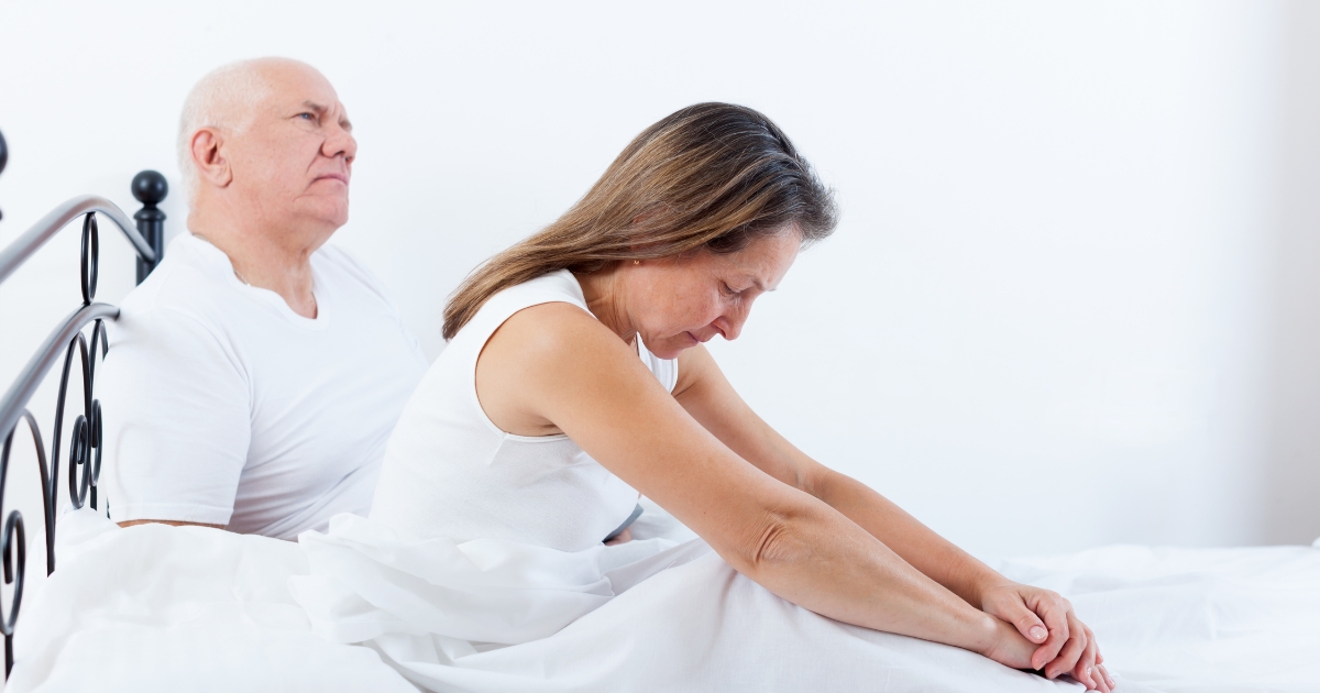 Older couple sitting in bed with emotional distance — woman appears sad and withdrawn while man looks away, symbolizing intimacy struggles in mature relationships.