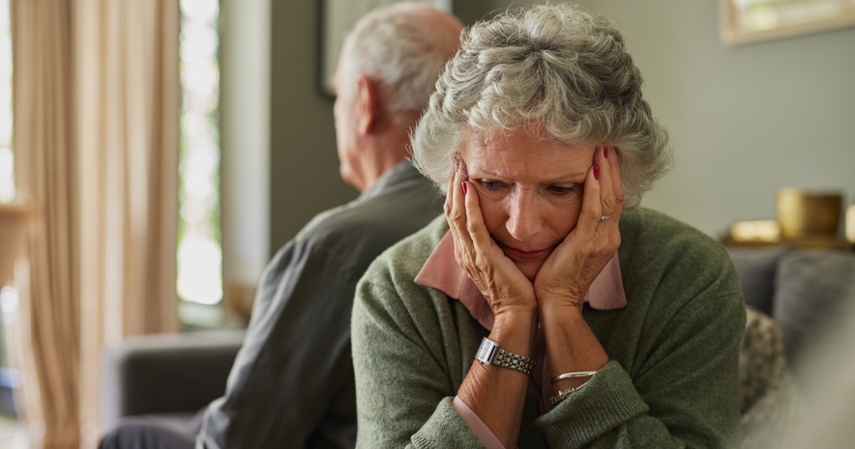 Older woman looking distressed and reflective, sitting apart from her partner, symbolizing emotional struggles and isolation during midlife divorce.