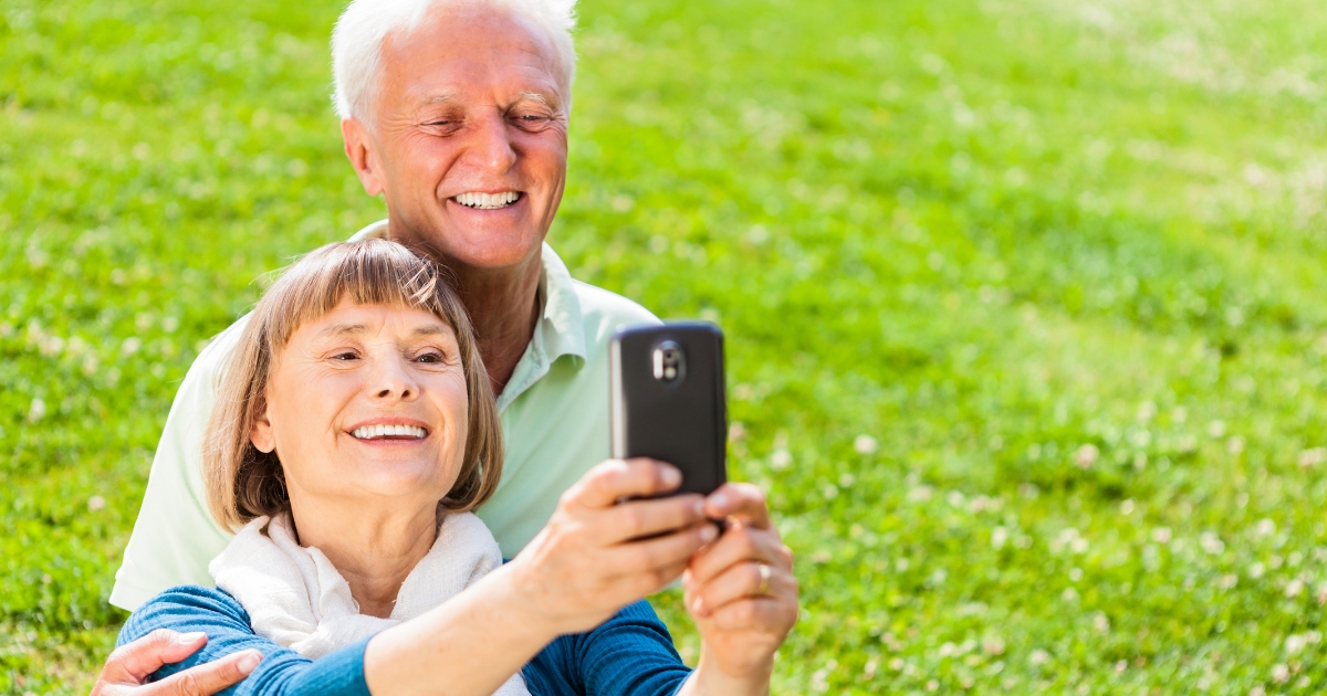 Smiling senior couple taking a selfie outdoors on a sunny day, enjoying a joyful moment together in nature, symbolizing love, connection, and shared experiences after 50.