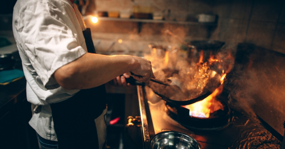Chef cooking with a wok over an open flame in a commercial kitchen, with flames and steam rising during high-heat stir-frying.