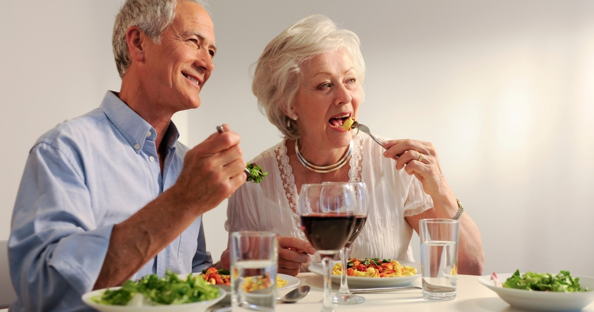 Smiling senior couple enjoying a meal together at a dining table, with plates of pasta, salad, glasses of water, and a glass of red wine.