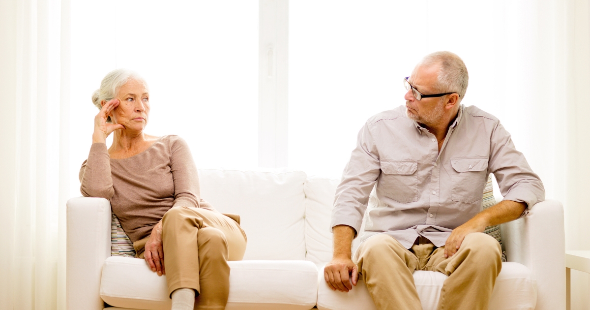 Older couple sitting apart on a couch with serious expressions, reflecting emotional distance and tension during a late-life divorce.