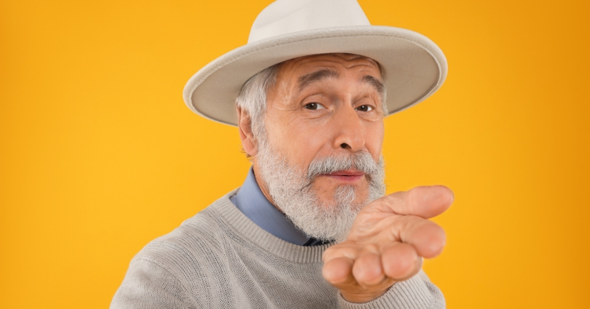 Stylish senior man with gray beard wearing a white hat and sweater blowing a kiss against a bright yellow background.