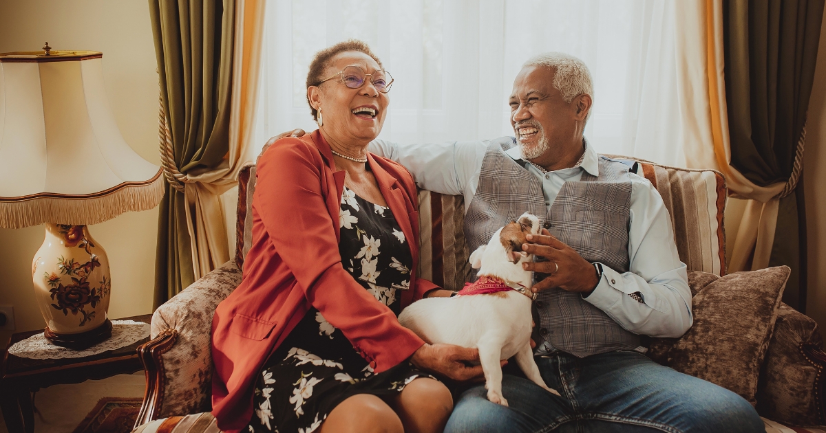 Joyful senior couple sitting on a couch at home, laughing together while holding their dog, capturing fun and companionship in later-life relationships.