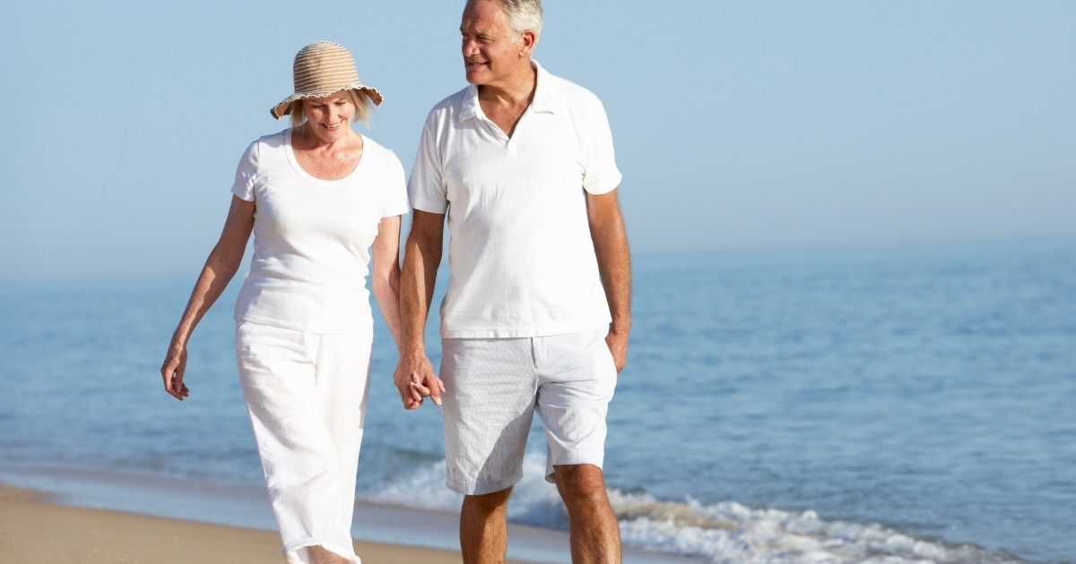 Happy senior couple holding hands and walking along the beach, dressed in white summer clothes, enjoying a peaceful day by the ocean.