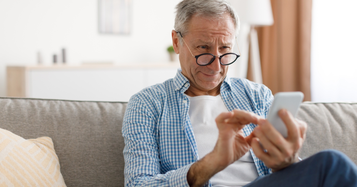 Senior man using smartphone while sitting on couch, exploring online dating apps for over 50s.