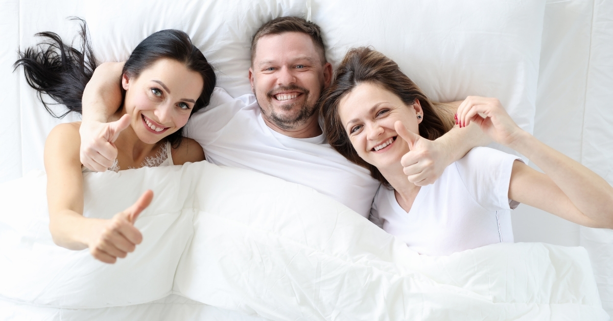 Smiling man in bed with two happy women giving thumbs up, representing a polyamorous relationship.