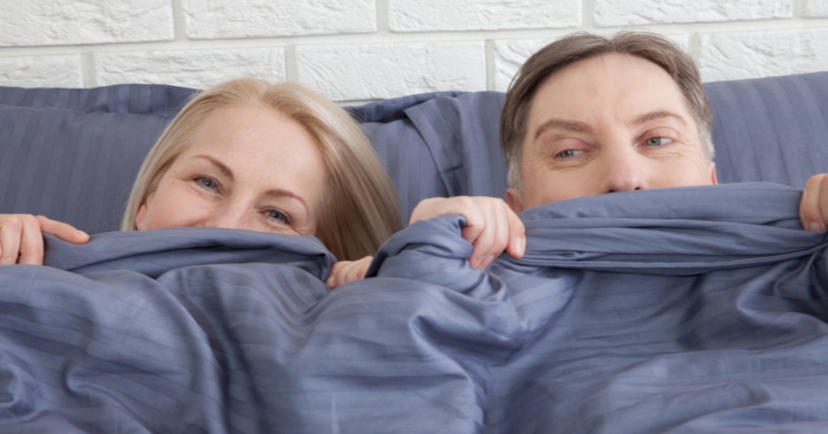 Playful mature couple in bed peeking from under blue covers, symbolizing intimacy and romance in midlife.