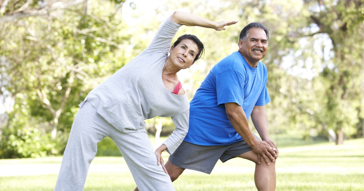 Older couple stretching together outdoors in a park, illustrating the benefits of physical activity and shared fitness for healthy relationships after 50.