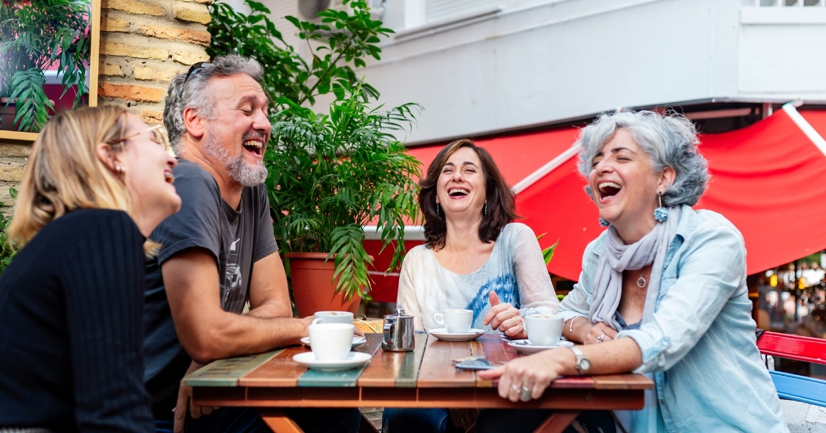 Group of older adults laughing and enjoying coffee together at an outdoor café, representing friendship, connection, and joy in later life.