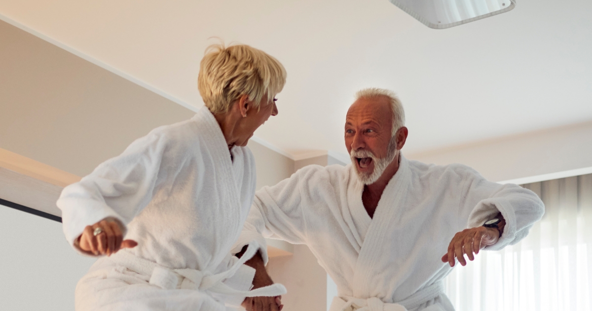 Joyful senior couple in white bathrobes laughing and jumping on a bed together in a bright room, enjoying a playful moment.