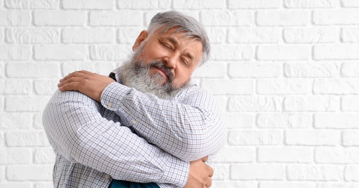 Mature man with gray hair and beard hugging himself with eyes closed, embracing self-love and emotional well-being after 50.