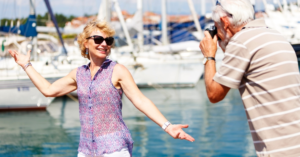 Senior couple enjoying a marina; woman poses playfully while man takes her photo, capturing joyful travel moments for mature adults near the water.