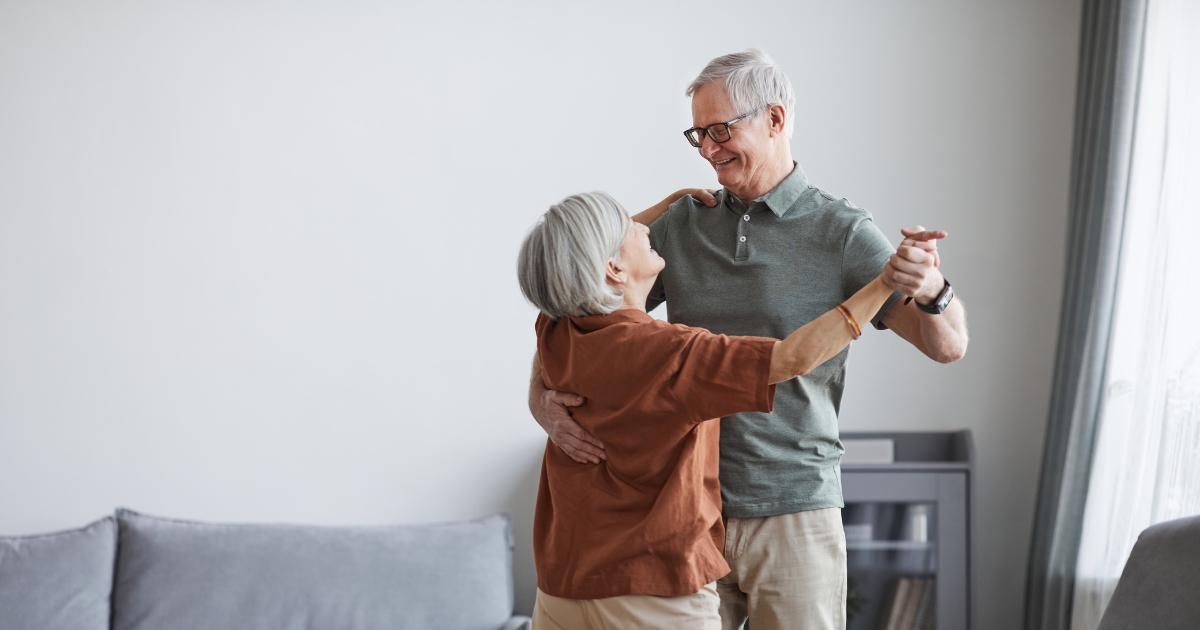 Senior couple dancing together in a cozy living room, smiling and enjoying a joyful moment, representing love and companionship in later life.