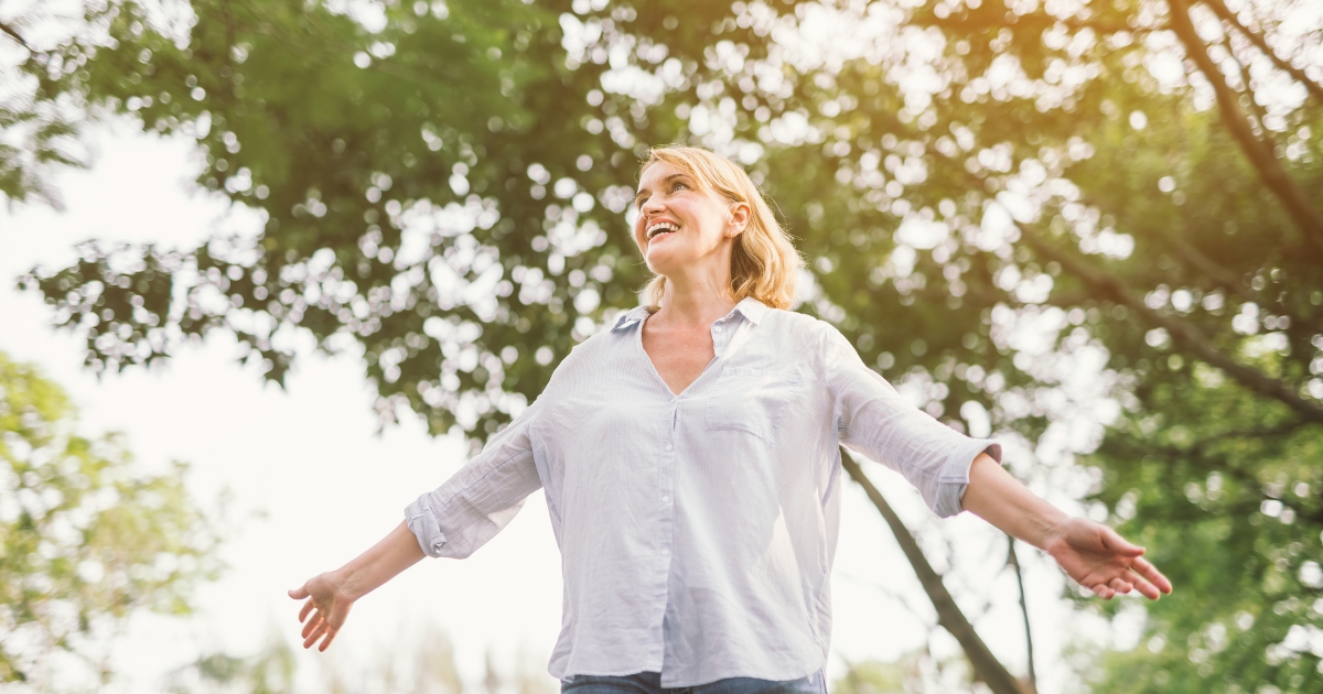 Joyful middle-aged woman standing outdoors with arms outstretched, smiling under the trees, symbolizing freedom, independence, and contentment in single life or personal growth.