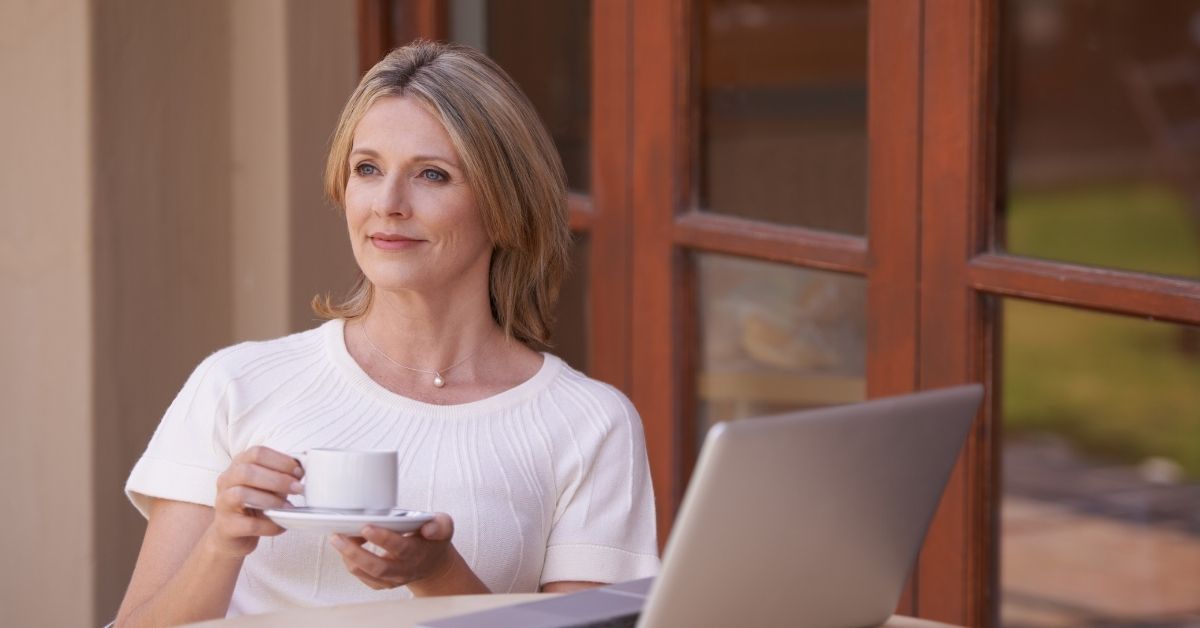 Smiling mature woman holding a coffee cup and looking thoughtful while sitting outside with a laptop, symbolizing self-reflection and exploring new relationship possibilities.