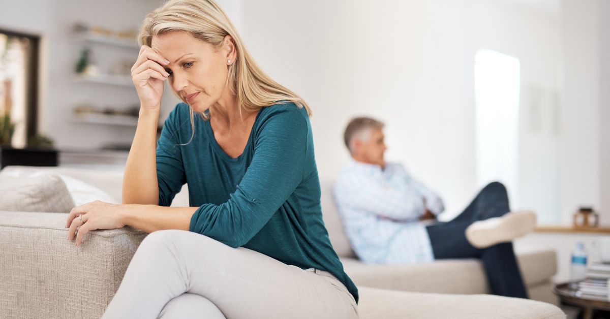 Frustrated woman sitting on couch with partner in background after a relationship conflict.