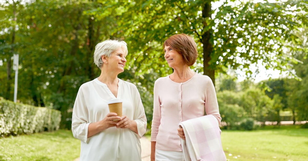 Two mature women walking outdoors in a park, smiling and talking while holding a coffee cup and a blanket—illustrating meaningful friendship and supportive conversations in later life.