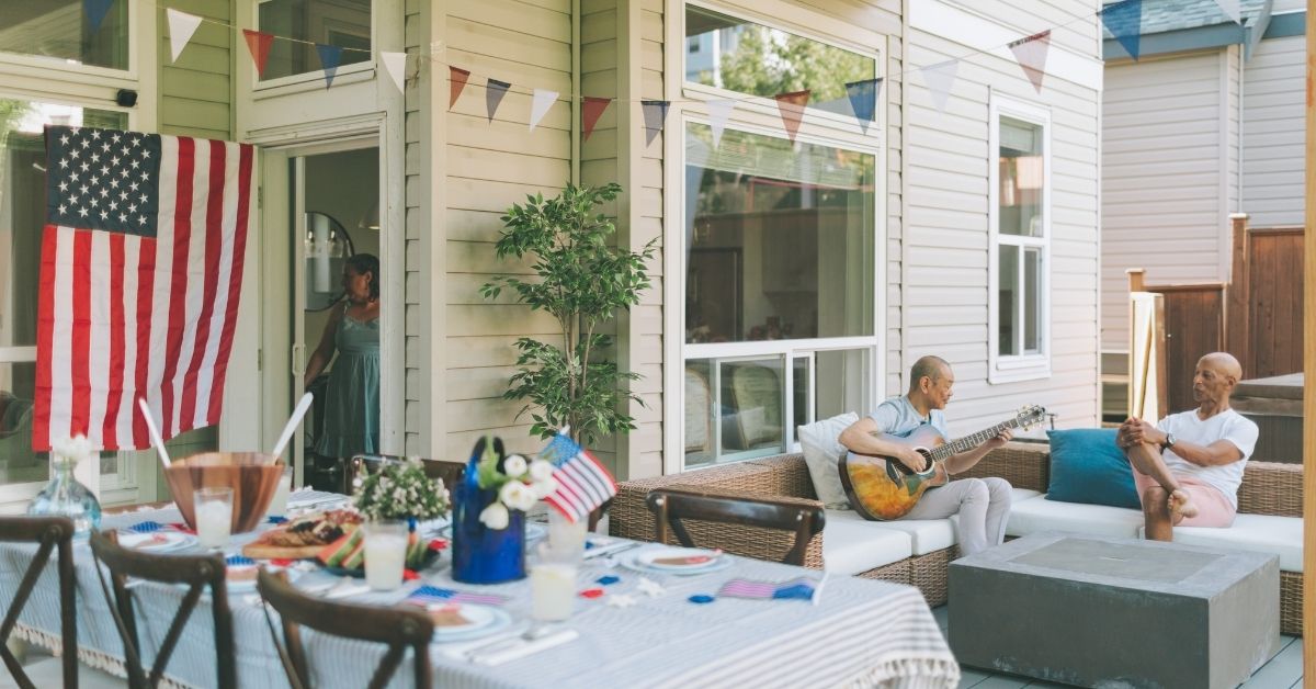 Backyard 4th of July celebration with a decorated patio, American flag, outdoor dining setup, and two men relaxing while one plays guitar, creating a festive atmosphere.