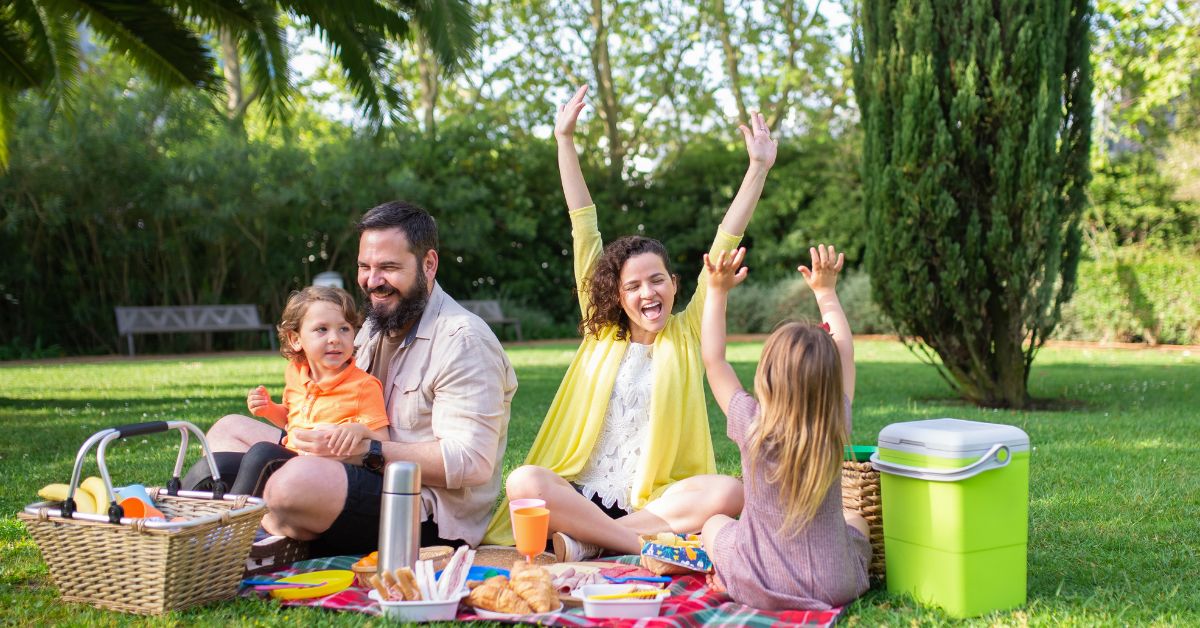 Happy family enjoying a picnic in a park, with two adults and two children sitting on a blanket surrounded by food and picnic items.